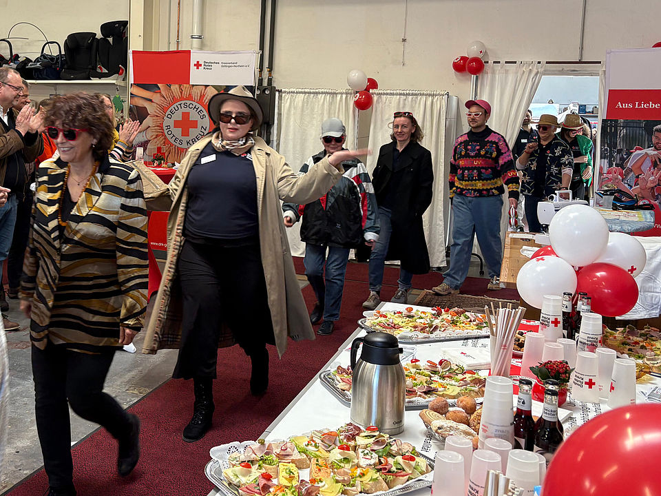 2026_03_03_Spendenzentrum_EInweihung_Modenschau.jpg Teilnehmende der Modenschau bewegen sich über den festlich dekorierten Teppichweg im DRK‑Spendenzentrum Göttingen. Im Vordergrund steht ein Buffet mit belegten Brötchen und Getränken, im Hintergrund findet die Modenschau statt