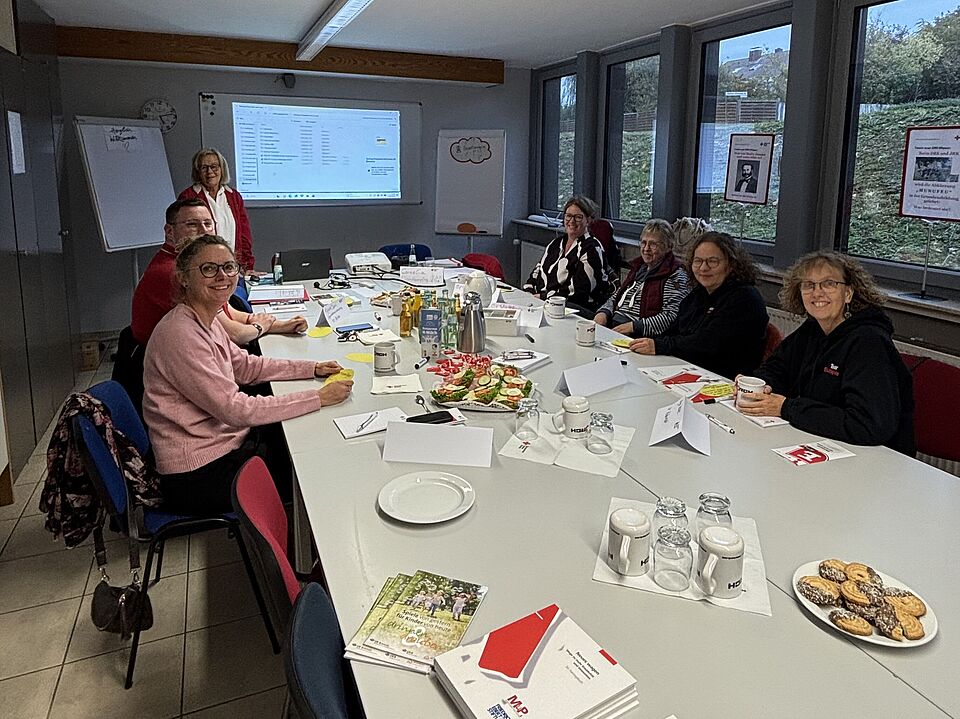 2026_10_23_DRK_Workshop_in_Northeim__5_.jpeg Die Teilnehmenden sitzen gemeinsam an einem Konferenztisch mit Schulungsunterlagen, Moderationsmaterial und Verpflegung. Im Hintergrund sind eine Präsentationsfläche und Pinnwände zu sehen.