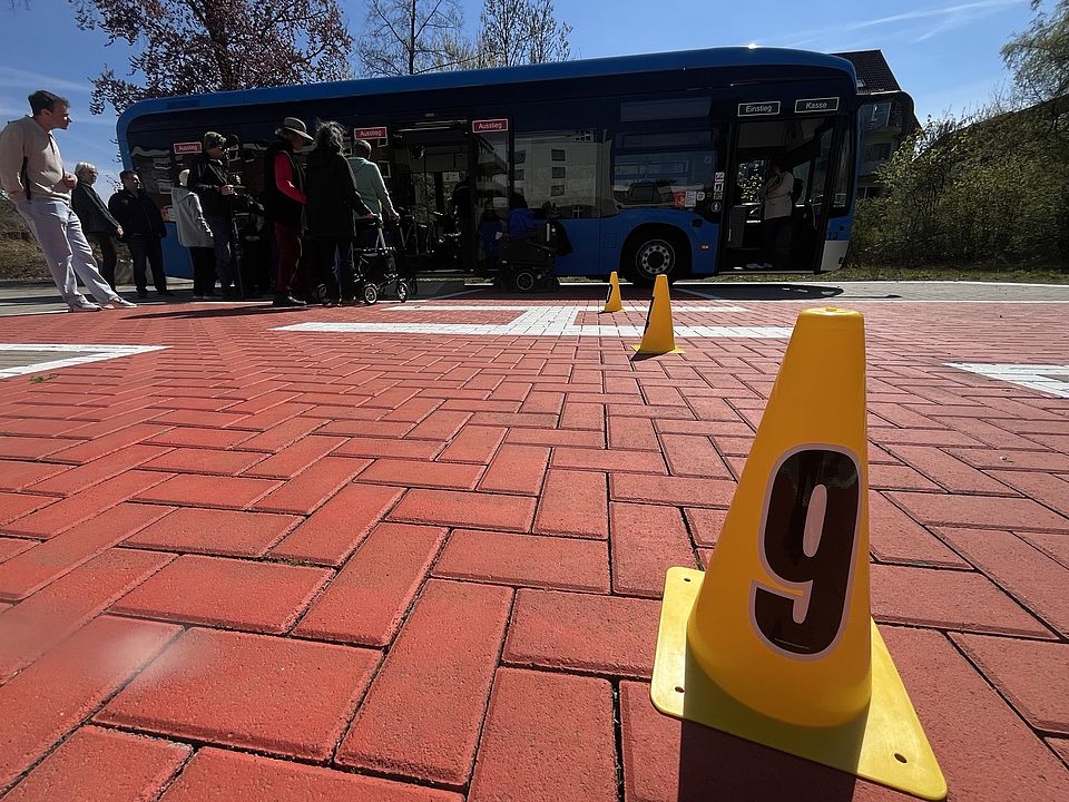 Teilnehmende am Stadtbustraining des DRK-Ortsvereins Göttingen in Kooperation mit dem SC Hainberg. Teilnehmende üben über eine Rampe das Einsteigen in den Bus mit einem Rollator, im Vordergrund ein Hütchen mit der Nummer 9 darauf auf einem großen Steinplatz