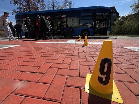 Teilnehmende am Stadtbustraining des DRK-Ortsvereins Göttingen in Kooperation mit dem SC Hainberg. Teilnehmende üben über eine Rampe das Einsteigen in den Bus mit einem Rollator, im Vordergrund ein Hütchen mit der Nummer 9 darauf auf einem großen Steinplatz