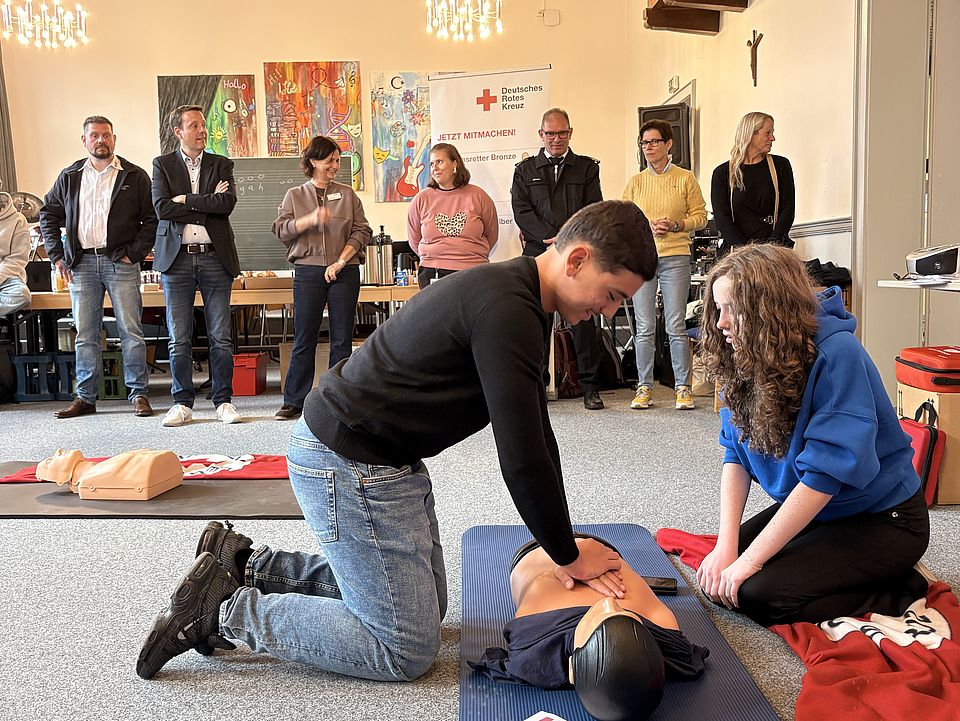 2026_09_21_Schüler_innen_üben_Herz-Lungen_Wiederbelebung_und_geladene_Gäste.jpg In einem Klassenraum trainieren zwei Schüler*innen die Herz-Lungen-Wiederbelebung an einer Übungspuppe. Im Hintergrund verfolgen mehrere geladene Gäste die Übung. Die Szene zeigt eine praktische Ausbildungseinheit im Rahmen der Woche der Wiederbelebung an der Bonifatiusschule in Göttingen