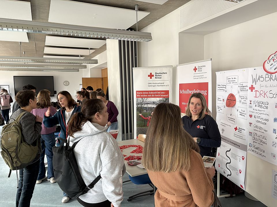 2026_02_25_Messe_Durchstarten_in_der_Sozialpägagogik_VHS__11_.JPEG In einem hellen Raum der VHS Göttingen Osterode stehen mehrere Personen an einem Informationsstand des Deutschen Roten Kreuzes (DRK‑Kreisverband Göttingen‑Northeim e. V.) auf der Jobmesse „Durchstarten in der Sozialpädagogik“. Rechts im Bild steht eine Person in DRK‑Dienstkleidung, die als hauptamtliche Mitarbeiterin tätig ist, hinter dem Stand und spricht mit mehreren Besuchenden. Auf dem Tisch vor ihr liegen Informationsflyer sowie zahlreiche kleine rote Kunststoffherzen, die dekorativ zu einer Form ausgelegt sind. Hinter dem Stand stehen zwei große DRK‑Roll‑up‑Banner mit Texten und Bildern zu pädagogischen Tätigkeitsfeldern, unter anderem zur Schulbegleitung. Links im Bild bewegen sich mehrere Besucherinnen und Besucher durch den Raum, einige mit Rucksäcken oder Taschen. Im Hintergrund sind weitere Stände und Menschen zu sehen, die Gespräche führen oder Materialien ansehen. Der Raum ist modern gestaltet, mit großen Fenstern und einer strukturierten Deckenbeleuchtung.