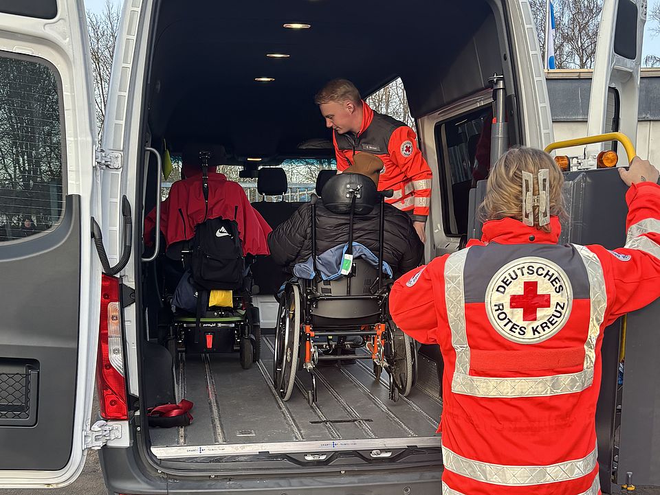 Man sieht einen Einsatzwagen der DRK Bereitschaft von Hinten, mit offener Heckklappe. Zwei Mitarbeitende der Bereitschaft helfen zwei Personen im Rollstuhl in das Fahrzeug, beide Personen im Rollstuhl sind bereits im Fahrzeug