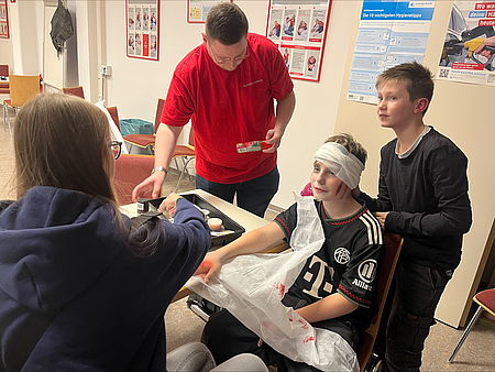 Zwei Kinder werden von zwei JRK Kolleg*nnen betreut. Der eine Junge steht hinter dem anderen, der einen Kopfverband trägt und am Arm verarztet wird