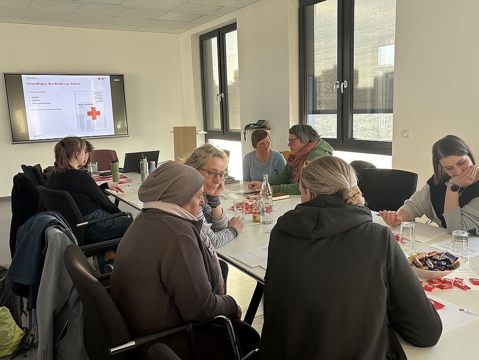 Ein heller Konferenzraum des Deutschen Roten Kreuzes in der Zimmermannstraße 4 in Göttingen. Mehrere Personen sitzen an einem großen rechteckigen Tisch und nehmen an einer Einführungsveranstaltung für neue Schulbegleiterinnen des FUD im DRK‑Kreisverband Göttingen‑Northeim e. V. teil. Auf einem großen Bildschirm an der Stirnseite des Raums wird eine Präsentationsfolie zum Thema „Grundlagen der Schulbegleitung“ angezeigt. Die Teilnehmenden sprechen miteinander, lesen Unterlagen oder schreiben Notizen. Auf dem Tisch liegen verschiedene Schulungsmaterialien, DRK‑Infobroschüren, kleine rote Give‑aways, Getränke sowie eine Schale mit Süßigkeiten. Durch große Fenster fällt viel Tageslicht in den Raum. Die Schulung wird von Alexandra Jäger geleitet.