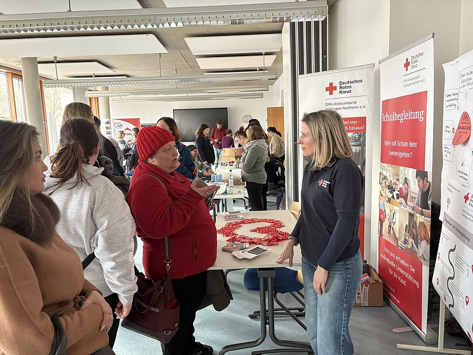 2026_02_25_Messe_Durchstarten_in_der_Sozialpägagogik_VHS__9_.JPEG In einem großen, hellen Raum der VHS Göttingen Osterode findet die Jobmesse „Durchstarten in der Sozialpädagogik“ statt. Viele Menschen bewegen sich zwischen mehreren Informationsständen und stehen in kleinen Gruppen zusammen. Im Vordergrund befindet sich ein Stand des Deutschen Roten Kreuzes (DRK‑Kreisverband Göttingen‑Northeim e. V.). Auf dem Tisch liegen Infomaterialien, Flyer sowie eine lange Kette aus roten Kunststoffherzen. Hinter dem Tisch steht DRK-Mitarbeiterin Bettina Sachreiter-Rund in dunkler DRK‑Dienstkleidung und spricht mit einer besuchenden Person. Rechts neben dem Tisch stehen zwei große Roll‑up‑Banner des DRK. Einer zeigt den Titel „Schulbegleitung“ und enthält mehrere Fotos sowie erklärende Texte zum Tätigkeitsfeld. Links und im Hintergrund sind weitere Stände anderer Organisationen zu sehen, an denen sich Besucherinnen und Besucher informieren. Durch die großen Fenster auf der linken Seite fällt Tageslicht in den Raum und beleuchtet die gesamte Messeszene.