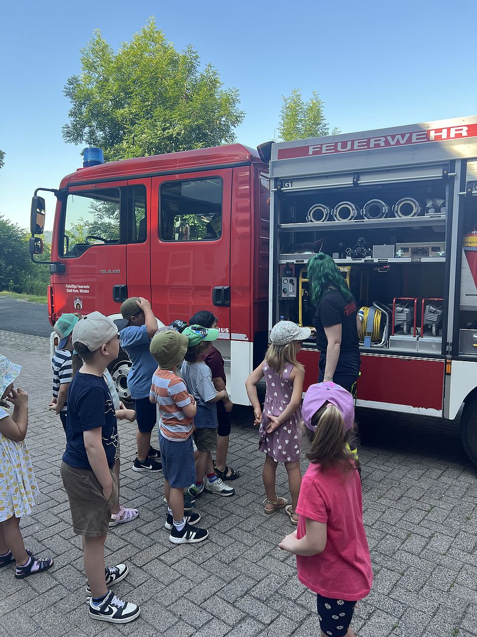Mehrere Kita-Kinder stehen draußen und bekommen von einer Feuerwehrfrau das Feuerwehrauto mit dem gesamten Zubehör und Innenleben erklärt