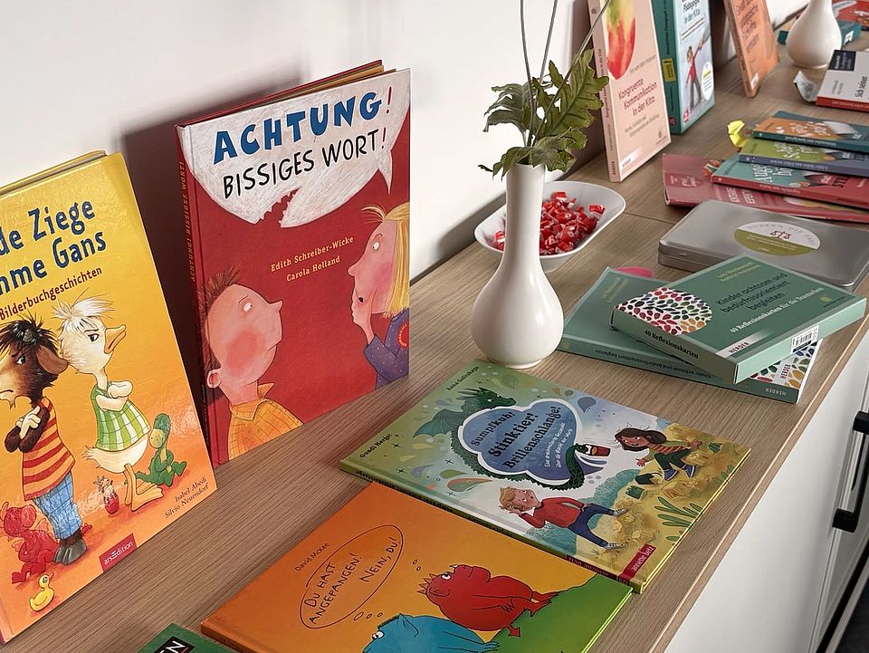 2026_07_07_Workshop_Sprache_und_Adultismus_für_pädagogische_Fachkräfte__24_.jpeg Auf einem langen Tisch stehen Kinderbücher und Fachmaterialien zum Thema Sprache und Verhalten, darunter „Achtung! Bissiges Wort!“ sowie weitere Bilderbücher. Eine Vase mit roten Blumen und kleine Materialien ergänzen die Präsentation für die interne Fortbildung des DRK-Kreisverbands Göttingen‑Northeim e. V.