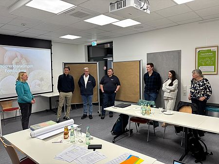 In einem Seminarraum des DRK-Kreisverbands Göttingen‑Northeim stehen mehrere Teilnehmende einer Erste‑Hilfe‑Fortbildung im Halbkreis. Eine Referentin spricht zur Gruppe, während hinter ihr eine Präsentationsfolie mit dem Titel „Kindererkrankungen – Fortbildung“ auf der Leinwand zu sehen ist. Die Personen hören aufmerksam zu. Im Raum befinden sich Tische mit Getränkeflaschen, Unterlagen und Materialien für die Schulung. Die Fortbildung findet im Rahmen einer praxisnahen Weiterbildung für Erste‑Hilfe‑Ausbilder*innen statt, die von Markus Schiffer, Tessa Lehmann und Niklas Benndorf gestaltet wird. Der Raum ist gut beleuchtet, neutral eingerichtet und mit Moderationsmaterial ausgestattet.