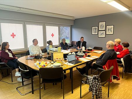 An einem Tisch sitzen ehrenamtliche Mitglieder des DRK-Kreisverbandes Göttingen Northeim anlässlich eines Austauschworkshops zur Blutspende. Auf dem Tisch stehen Getränke und Arbeitsmaterialien und im Hintergrund ist ein rotes Kreuz am Fenster