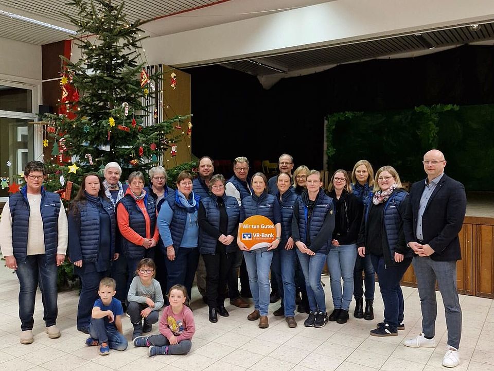 2025_12_Spendenübergabe_VR_Bank_OV_Drüber-Sülbeck.jpg In einem festlich dekorierten Innenraum steht eine größere Gruppe von Personen vor einem geschmückten Weihnachtsbaum. Die Gruppe besteht aus Ehrenamtlichen des DRK‑Ortsvereins Drüber‑Sülbeck, die überwiegend dunkelblaue, einheitliche Westen tragen. In der Mitte hält eine Person ein großes, orangefarbenes Schild mit dem Logo der VR‑Bank und der Aufschrift „Wir tun Gutes“. Im Vordergrund sitzen mehrere Kinder auf dem Boden. Im Hintergrund sind eine Bühne mit dunklem Vorhang sowie Raumbeleuchtung und Decke mit sichtbaren Paneelen zu erkennen. Das Foto dokumentiert die Spendenübergabe der VR‑Bank (1.000 Euro), mit der die neuen Westen für die ehrenamtliche Arbeit – unter anderem bei Blutspende‑Einsätzen – finanziert wurden