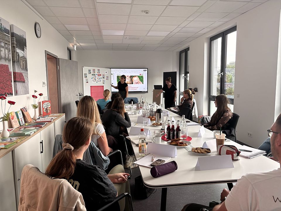 2026_07_07_Workshop_Sprache_und_Adultismus_für_pädagogische_Fachkräfte__12_.jpeg Interne DRK‑Fortbildung zu Sprache und Adultismus: pädagogische Fachkräfte im Workshop in Göttingen. Teilnehmende sitzen an einem Konferenztisch mit Arbeitsmaterialien, während vorne Inhalte präsentiert werden. Auf einem Nebentisch liegen Bücher und Materialien zum Thema Sprache im Kita-Alltag.