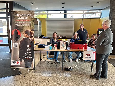 Es sind Ehrenamtliche des DRK-Kreisverbandes Göttingen-Northeim e. V. zu sehen, wie sie an der Anmeldung um einen Tisch bei der Blutspende in Dassel stehen. Im Vordergrund sieht man einen Aufsteller des DRK