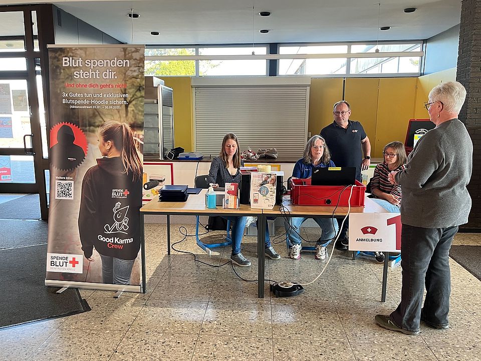 Es sind Ehrenamtliche des DRK-Kreisverbandes Göttingen-Northeim e. V. zu sehen, wie sie an der Anmeldung um einen Tisch bei der Blutspende in Dassel stehen. Im Vordergrund sieht man einen Aufsteller des DRK