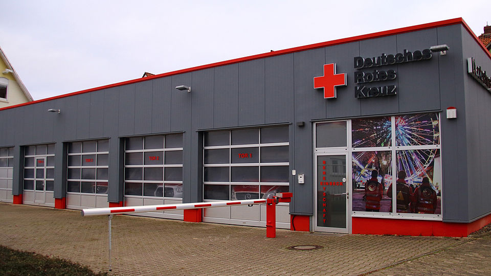 2026_01_29_Umzug_Bereitschaft_Einbeck__3_.jpg Man sieht die DRK-Bereitschaft Einbeck, ein graues flaches Gebäude mit dem DRK Logo rechts oben über der Tür und einer rot-weißen Schranke davor