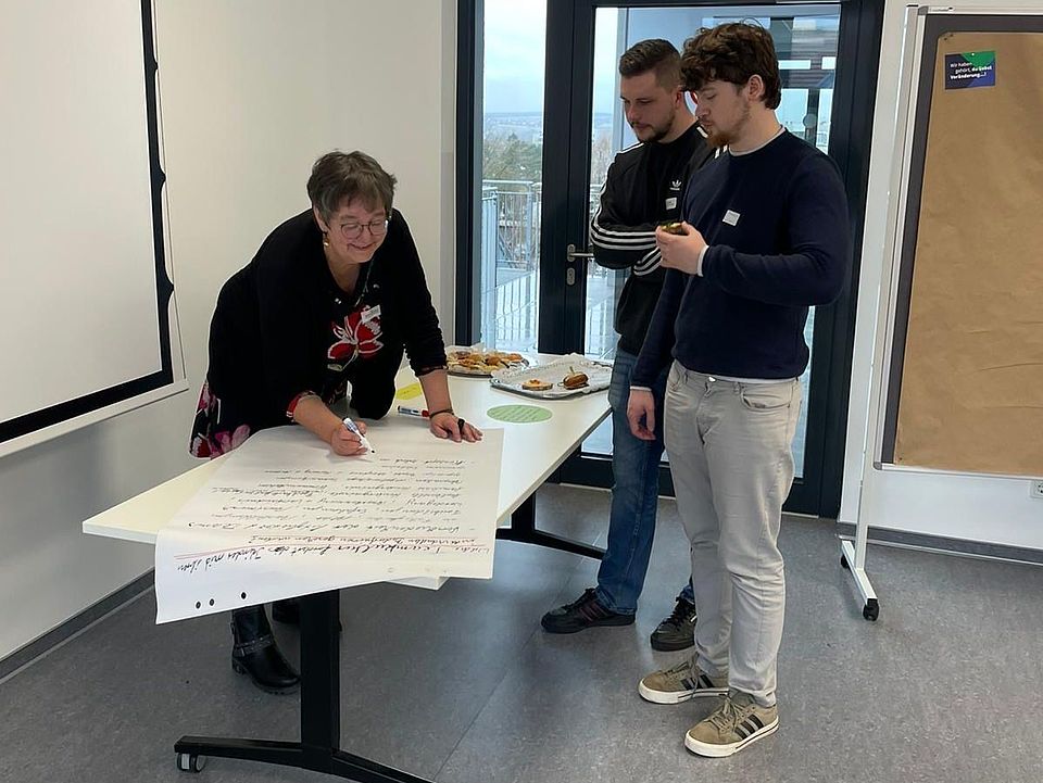 In einem Konferenzraum des DRK‑Kreisverbandes Göttingen‑Northeim e. V. steht eine Person an einem Tisch und schreibt auf ein großformatiges Plakatpapier, das für die Dokumentation der World‑Café‑Ergebnisse genutzt wird. Neben ihr stehen zwei weitere Personen und betrachten den Text. Hinter ihnen befindet sich eine Glastür mit Blick nach draußen sowie ein kleiner Tisch mit Tellern und Snacks, die zum Auftakt des Fachtags gereicht wurden. Rechts steht eine mobile Pinnwand mit braunem Papier, die für weitere Workshop‑Notizen vorgesehen ist. Die Szene zeigt einen Arbeitsmoment während der vertiefenden Reflexion zur Umsetzung inklusiver Schutzkonzepte.