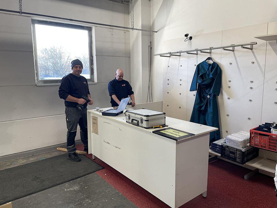 Zwei DRK Kollegen stehen in der Halle des neuen Spendenzentrums und montieren eine Tresen. An der Rückwand ist bereits ein blaues Damenkleid  zu sehen, was an einer Garderobe hängt.