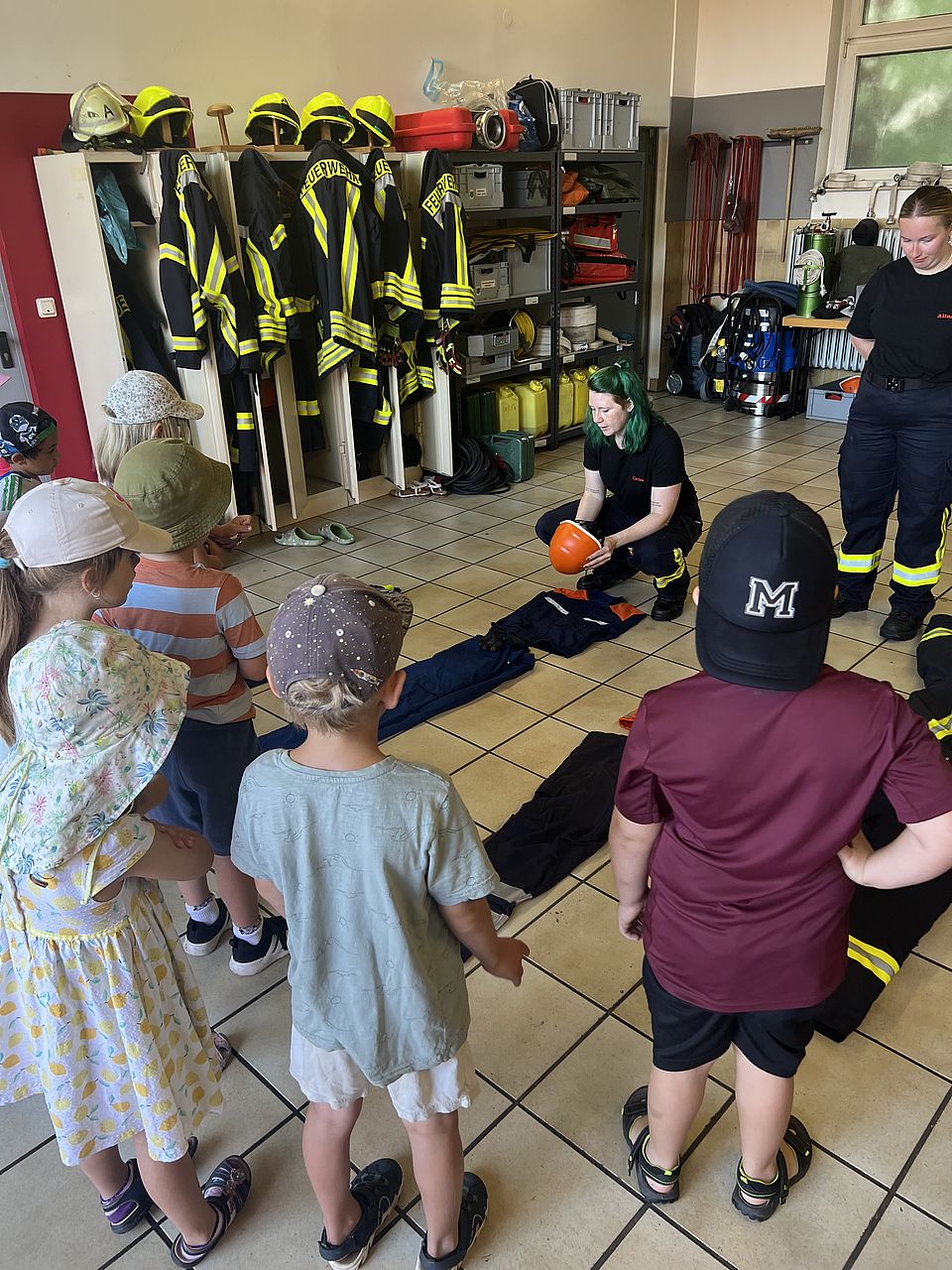 Mehrer Kita-Kinder stehen in einem Feuerwehr-Gerätehaus und beobachten zwe Feuerwehrfrauen, die ihnen die Uniform erklären. Die unterschiedlichen Hosen liegen auf dem Boden und ein Feuerwehrfrau häkt einen orangefarbenen Helm in der Hand. Im Hintergrund sind Regale und Schränke zu sehen, in denen das gesamte Equipment aufbewahrt wird.