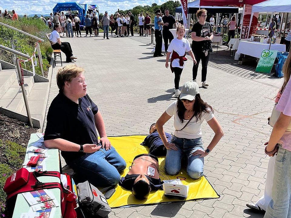 2026_06_06_1._Gesundheitstag_Ebergötzen_mit_OV_und_Erste_Hilfe__23_.jpg Mehrere Übungsmatten mit Reanimationspuppen liegen vor einem Stand mit Flyern des DRK. DRK‑Helfende zeigen Wiederbelebungsmaßnahmen, während Besuchende vorbeigehen und zuschauen.