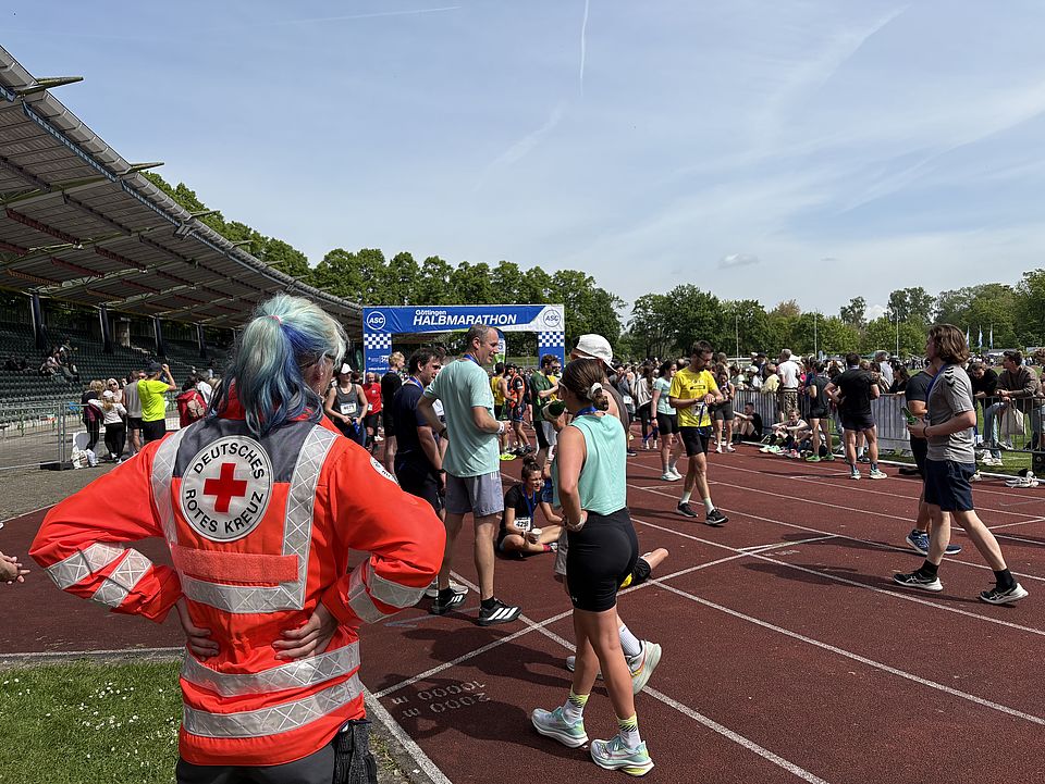 2026_5_10_Göttinger_Halbmarathon__11_.jpg Einsatz der DRK-Bereitschaft im Sanitätsdienst beim Göttinger Halbmarathon: Bereitschaftsmitglied von hinten mit DRK-Jacke, im Hintergrund der Start und Zielbereich des Halbmarathons im Jahnstadion sowie Läufer*innen