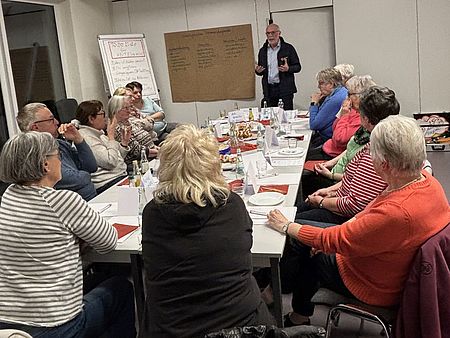 DRK-Ehrenamtlicher Herr Gniewosz steht vor einer großen, handschriftlich beschriebenen Papierwand und erklärt ein Organigramm zum möglichen Zusammenschluss mehrerer DRK‑Ortsvereine. Mehrere Ehrenamtliche sitzen am Tisch, hören zu und diskutieren