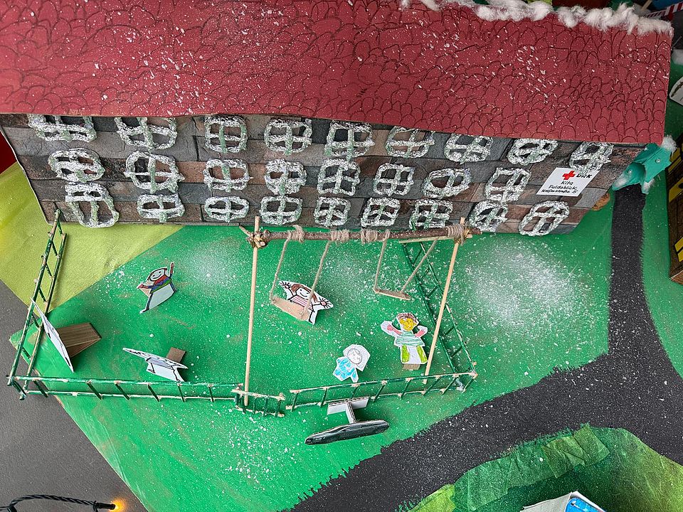 Aus Pappe gebastelt zu shehen ist das Gebäude der Kita Fuldablick mit Figuren, die auf einer grünen Fläche vor dme Haus im Gartenspielen