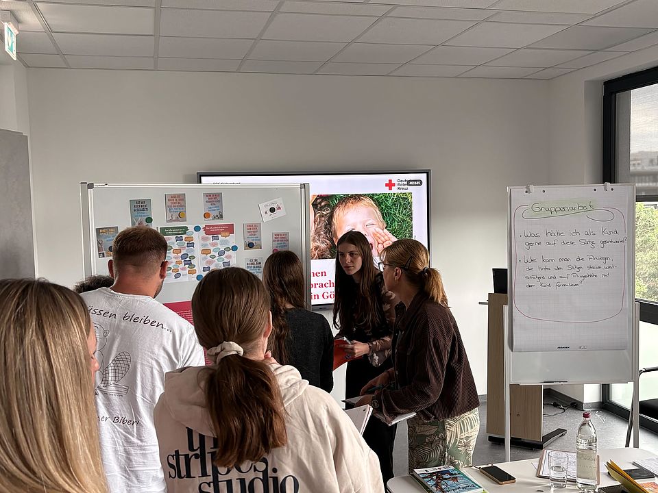 2026_07_07_Workshop_Sprache_und_Adultismus_für_pädagogische_Fachkräfte__20_.jpeg Teilnehmende stehen gemeinsam vor einer Pinnwand mit Plakaten und diskutieren Arbeitsaufgaben. Auf einem Flipchart stehen Fragen zur Reflexion von Sprache und Wirkung im Umgang mit Kindern.