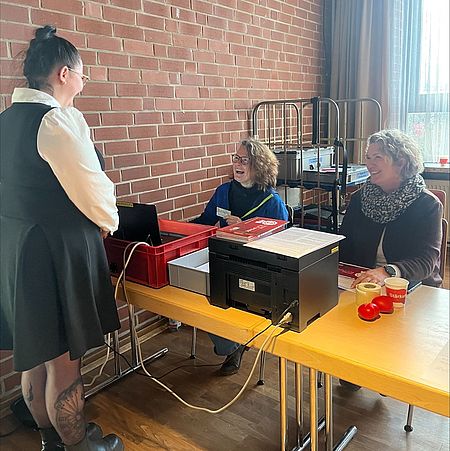 In einem Raum mit roter Ziegelwand und großem Fenster steht eine DRK-Ehrenamtliche an einem langen Tisch, an dem zwei weitere Ehrenamtliche sitzen. Auf dem Tisch stehen mehrere Laptops, Kisten mit Formularen, ein Drucker sowie Becher mit Getränken. Die beiden sitzenden Personen arbeiten an der Anmeldung oder Registrierung für eine Blutspendeaktion in Moringen. Hinter dem Tisch stehen Metallwagen mit weiterem Material. Die Szene zeigt den Anmeldebereich während einer Blutspendeveranstaltung in Moringen.