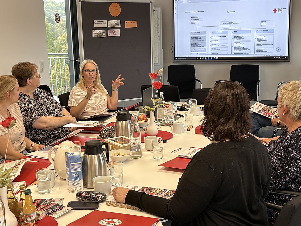 2026_10_01_Teamtag_Kita_Löwenhagen__11_.jpg Die Teilnehmenden sitzen gemeinsam an einem Besprechungstisch und beschäftigen sich mit Strukturen, Werten und Arbeitsweisen des DRK. Auf einem Bildschirm wird eine Präsentation des DRK-Kreisverbandes Göttingen-Northeim gezeigt.