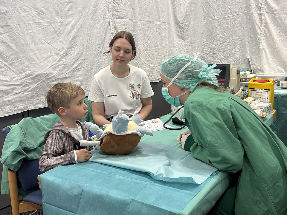 2026_06_01TBK_Teddybärkrankenhaus_UMG__Kita_Bovenden_11_.jpg Ein Kind sitzt vor einem Tisch mit einem Kuscheltier, während eine medizinisch gekleidete Person sich auf Augenhöhe dazu beugt. Eine weitere betreuende Person sitzt daneben und begleitet die Untersuchungssituation.