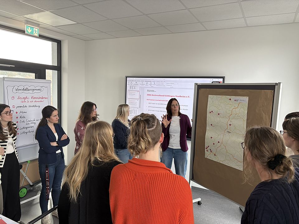 Teilnehmende eines Einführungstages des DRK-Kreisverbands Göttingen‑Northeim stehen gemeinsam vor einer Präsentationsfläche und einer großen Karte. DRK-Mitarbeitende Marion Laudenbach erklärt Inhalte zu Standorten und Strukturen des Verbandes
