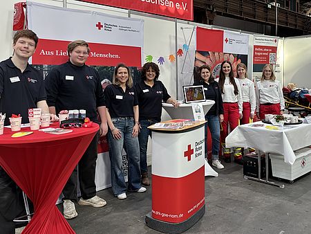 Gruppenaufnahme am DRK‑Messestand. Vorn sind mehrere Stehtische und Präsentationsflächen zu sehen, im Hintergrund Roll‑ups und Material zu Angeboten des Deutschen Roten Kreuzes