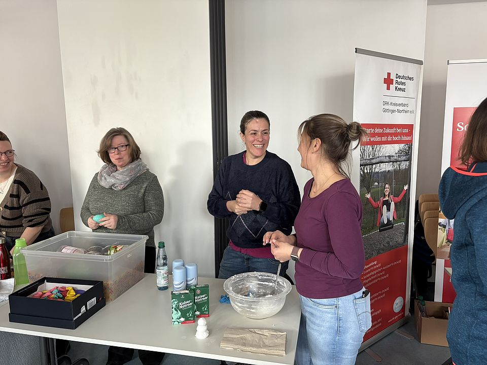 2026_02_25_Messe_Durchstarten_in_der_Sozialpägagogik_VHS__5_.JPEG An einem Tisch innerhalb eines Veranstaltungsraums stehen mehrere Personen zusammen und bereiten Materialien für eine Mitmach‑Aktion vor. Auf dem Tisch liegen verschiedene Gegenstände, darunter eine große transparente Kunststoffbox mit bunten Utensilien, Getränkeflaschen, mehrere Packungen Bastel‑ oder Experimentiermaterial sowie eine Schüssel mit einer weißen, schaumigen Mischung und Werkzeugen zur Verarbeitung. Hinter dem Tisch steht ein Roll‑up‑Banner des Deutschen Roten Kreuzes (DRK‑Kreisverband Göttingen‑Northeim e. V.), das Informationen zu pädagogischen Angeboten zeigt. Die Person auf der rechten Seite des Bildes ist eine Mitarbeitende einer DRK‑Kita und spricht mit einer der anderen anwesenden Personen. Der Raum ist hell und offen gestaltet, mit weißen Wänden und großen Fenstern, durch die Tageslicht einfällt.