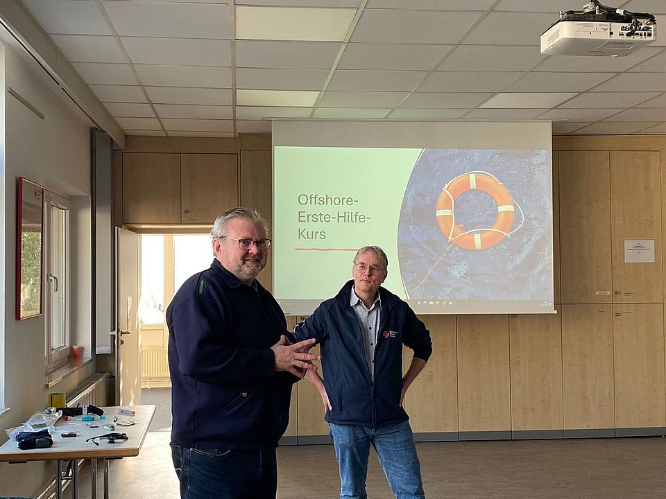 Zwei Personen, einer davon Markus Schiffer, stehen vor einer projizierten Präsentationsfolie mit dem Titel „Offshore-Erste-Hilfe-Kurs“. Auf dem Bildschirm ist ein Rettungsring über einem Meeresmotiv zu sehen. Der Raum ist mit Schulungsinventar wie Beamer, Tisch und medizinischen Übungsmaterialien ausgestattet.