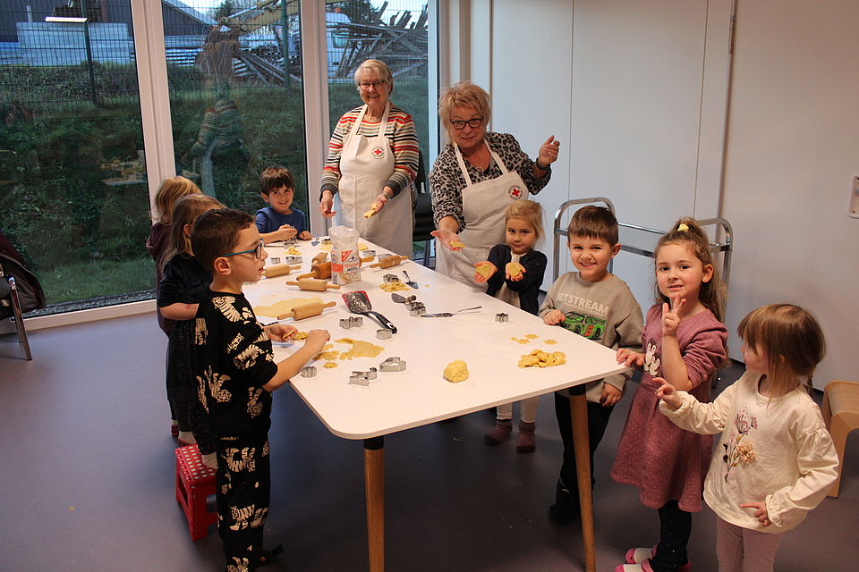 2025_12_04_Weihnachtsbacken Kordes-Kita Uslar Eine Gruppe an Kindern stehen um einen Tisch herum, sowie zwei Damen aus dem DRK-Ortsverein Uslar. Alle backen gemeinsam Kekse und lachen in die Kamera