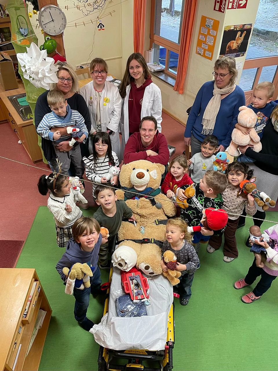 2025_11_24_Teddybärkrankenhaus__3 Ein Gruppenbild von Kindern aus der Kita Bovenden, Leiter Matthias Henne und Mitarbeitenden des Teddybärkrankenhauses, zusammen mit einem großen Teddybären, von oben fotografiert