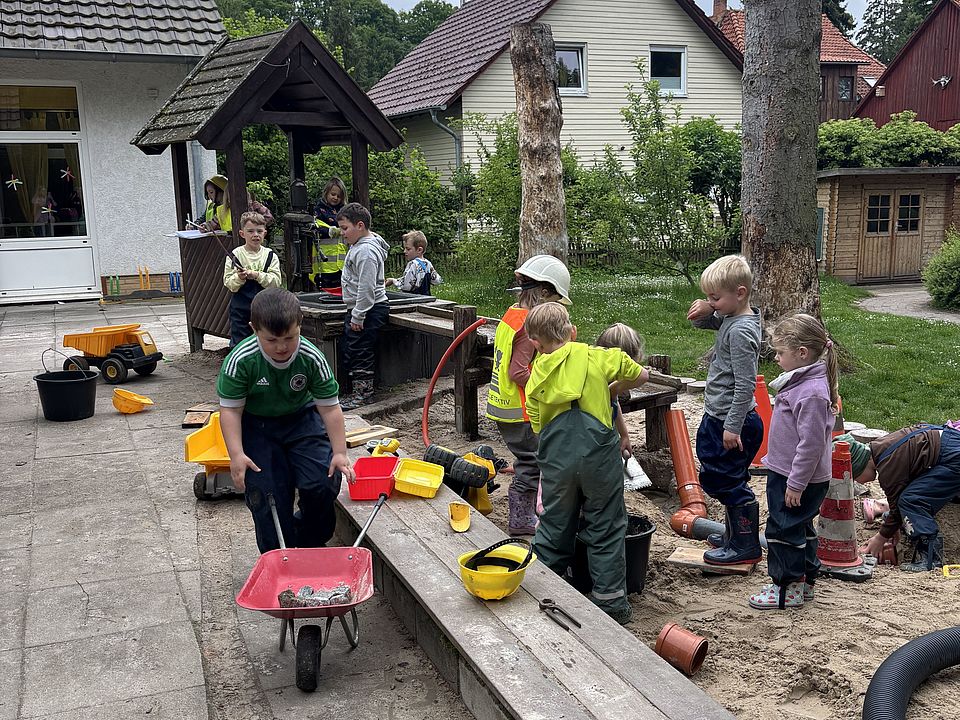2026_05_27_Baustelle.jpeg Mehrere Kinder spielen auf dem Kita‑Außengelände an einer improvisierten Baustelle. Eine Schubkarre, Eimer, Spielzeugfahrzeuge und Rohre liegen bereit, während die Kinder Sand und Steine bewegen.