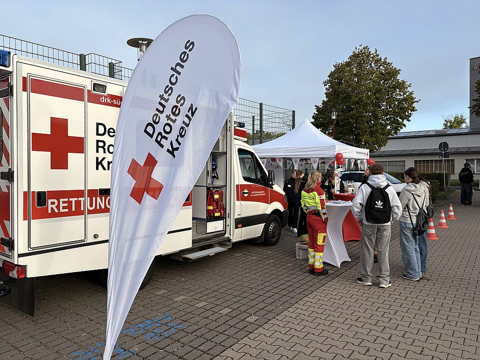 Schüler*innen der BBS Einbeck informieren sich am DRK-Stand auf der Einbecker Ausbildungsmesse, Beachflag des DRK und Rettungswagen mit Pavillion daneben