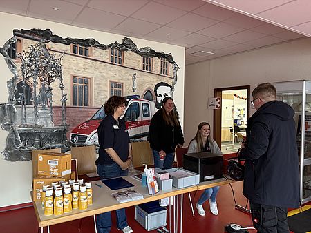Blutspendeaktion beim DRK-Kreisverband Göttingen-Northeim e. V., Anmeldetisch mit Mitarbeitenden und einem Spender bei der Registrierung vor einer Wand mit DRK-Motiv