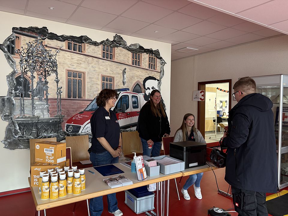 Blutspendeaktion beim DRK-Kreisverband Göttingen-Northeim e. V., Anmeldetisch mit Mitarbeitenden und einem Spender bei der Registrierung vor einer Wand mit DRK-Motiv