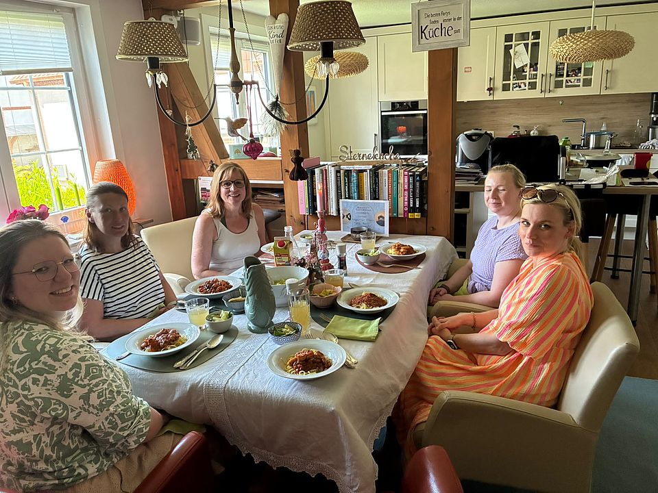2026_05_26_Teamtag_Kita_Bühren__3_.jpeg Team der Kita Bühren sitzt an gedecktem Tisch in heller Wohnküche und isst gemeinsam Pasta beim Teamtag.