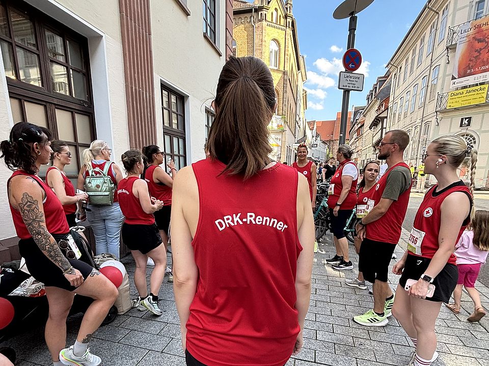 2026_07_01_Altstadt-Läuferinnen_vorher__7_.jpeg Teilnehmende in roten Shirts mit der Aufschrift „DRK-Renner“ stehen in einer Straße der Göttinger Innenstadt und bereiten sich auf den Altstadtlauf Göttingen 2026 vor.