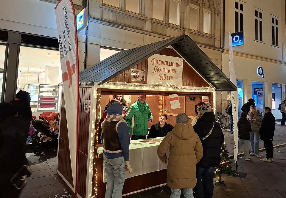 Freiwillig-in-Göttingen-Hütte am 5.12.25 auf dem Weihnachtsmarkt Die Freiwillig-in-Göttingen-Hütte mit zwei DRK Ehrenamtlichen und drei Interessierten, die sich am Stand informieren bei Dunkelheit