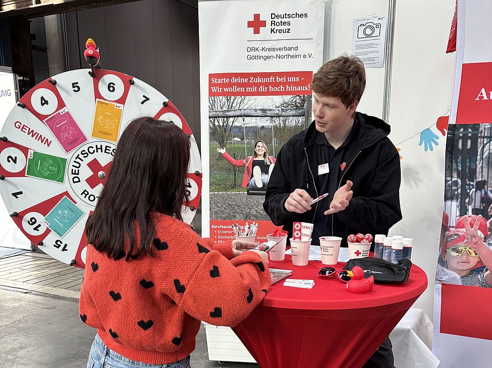 Stehtisch des DRK‑Kreisverbands Göttingen‑Northeim e. V. beim GöBit 2026. Ein DRK‑Mitarbeitender spricht mit einer Besucherin. Auf dem Tisch liegen Flyer, Becher und weitere Informationsmaterialien
