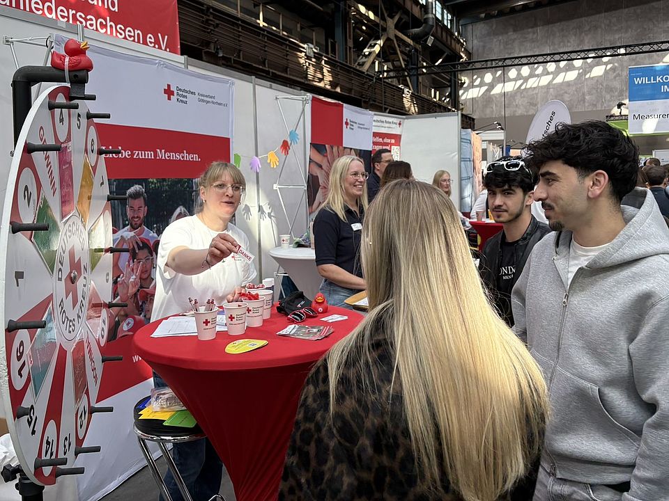 Belebter DRK‑Messestand beim GöBit 2026 in der Göttinger Lokhalle. Ein DRK‑Tisch mit Werbematerialien und einem Glücksrad ist sichtbar, während Besuchende mit Mitarbeitenden ins Gespräch kommen. Eine Mitarbeiterin reicht einer Besucherin einen Sticker