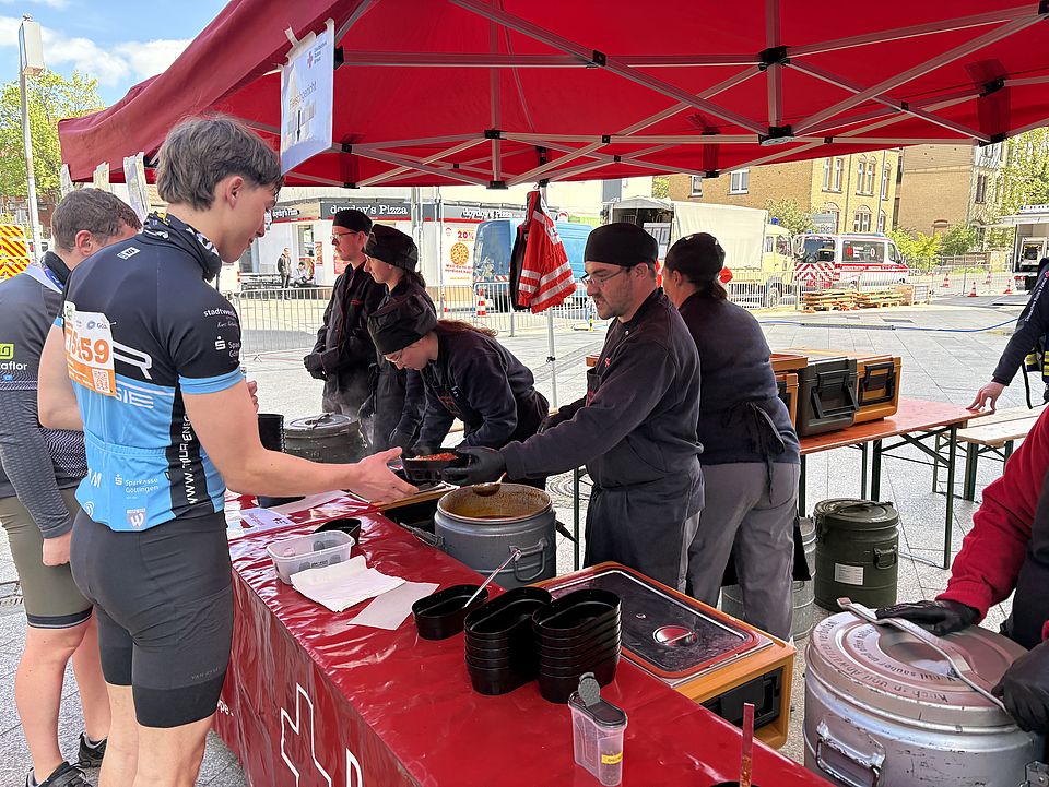 Helfer*innen und Mitglieder der DRK-Bereitschaft bei der Tour d'Energie 2026 in Göttingen beim Verpflegungsdienst. Ein Helfer gibt Essen an einen Radfahrer aus