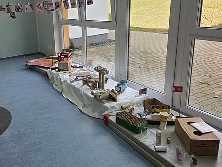 Ausstellung im Kita‑Flur mit mehreren von Kindern gebauten Konstruktionen aus Alltagsmaterialien wie Kartons, Rollen und Verpackungen. Die Werke stehen entlang großer Fenster auf Podesten