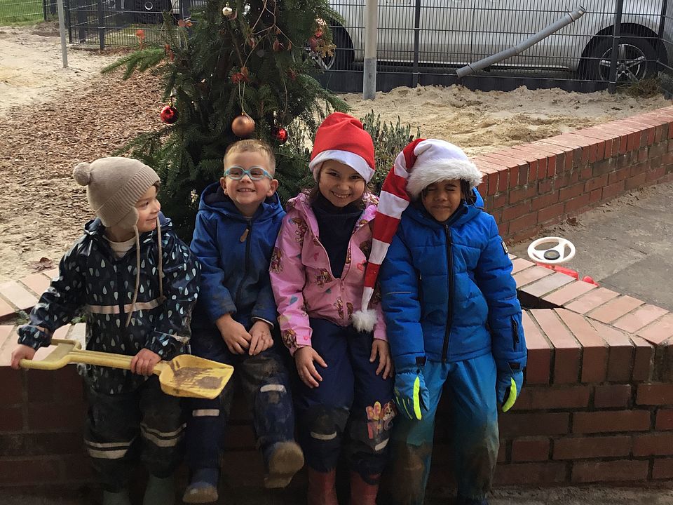 2025_12_11_Adventszeit_Kita_Wiershausen__1 Vier Kinder aus der Kita Wiershausen sitzen auf einer kleinen Mauer und lachen in die Kamera. Im Hintergrund ist ein Parkplatz und hinter den Kindern ein Weihnachtsbaum