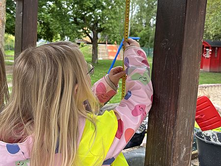 Ein Kind hält einen aufgeklappten Zollstock an eine Holzstrebe eines Spielgeräts und misst sorgfältig die Höhe. Im Hintergrund sind Bäume, Spielgeräte und das Kita‑Außengelände sichtbar.