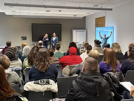 In einem großen Seminarraum sitzen zahlreiche Personen in Reihen von Stühlen und hören einem Vortrag zu. Vorne im Raum stehen zwei DRK-Mitarbeiterinnen nebeneinander und sprechen zur Gruppe. Hinter ihnen befinden sich ein großes Display, ein Flipchart sowie ein Tisch. Links am Rand steht eine weitere Person neben einem Fenster. Der Raum ist modern eingerichtet, mit einer Decke aus Akustikplatten und breiten Fenstern, durch die Tageslicht hereinfällt. Rechts im Hintergrund steht ein blaues Roll‑up‑Banner mit einem Motiv einer jubelnden Person und dem Schriftzug „Vielfalt – Kompetenz – Weiterbildung“. Die Szene zeigt einen Programmpunkt der Jobmesse „Durchstarten in der Sozialpädagogik“ an der VHS Göttingen Osterode.